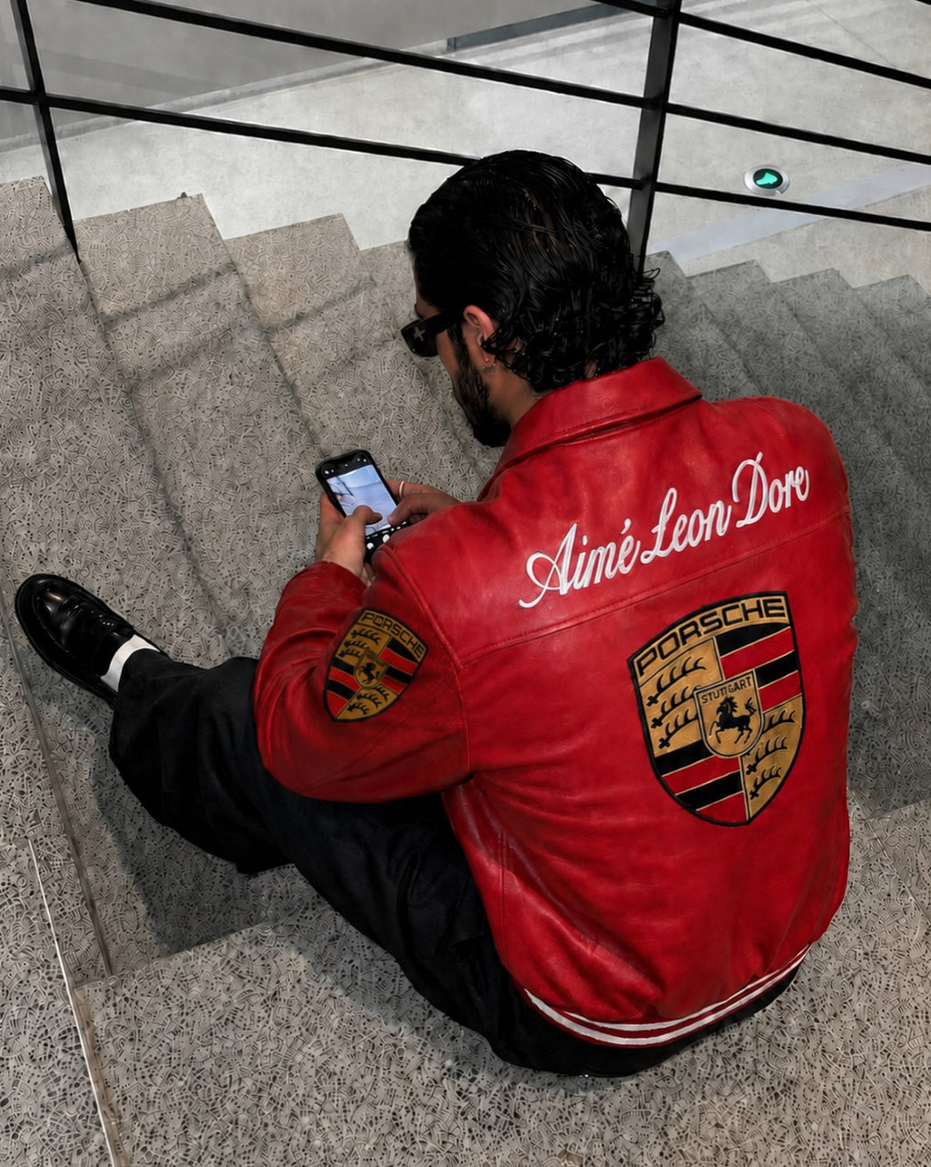 A man sitting on stone stairs wearing a red leather jacket with 'Amié Leon Dore' and a Porsche crest on the back, looking at his phone.