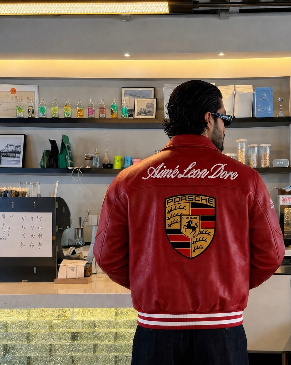 A man viewed from behind wearing a red leather jacket with 'Ambi Leon Dore' and a Porsche crest on the back, standing in a cafe setting.